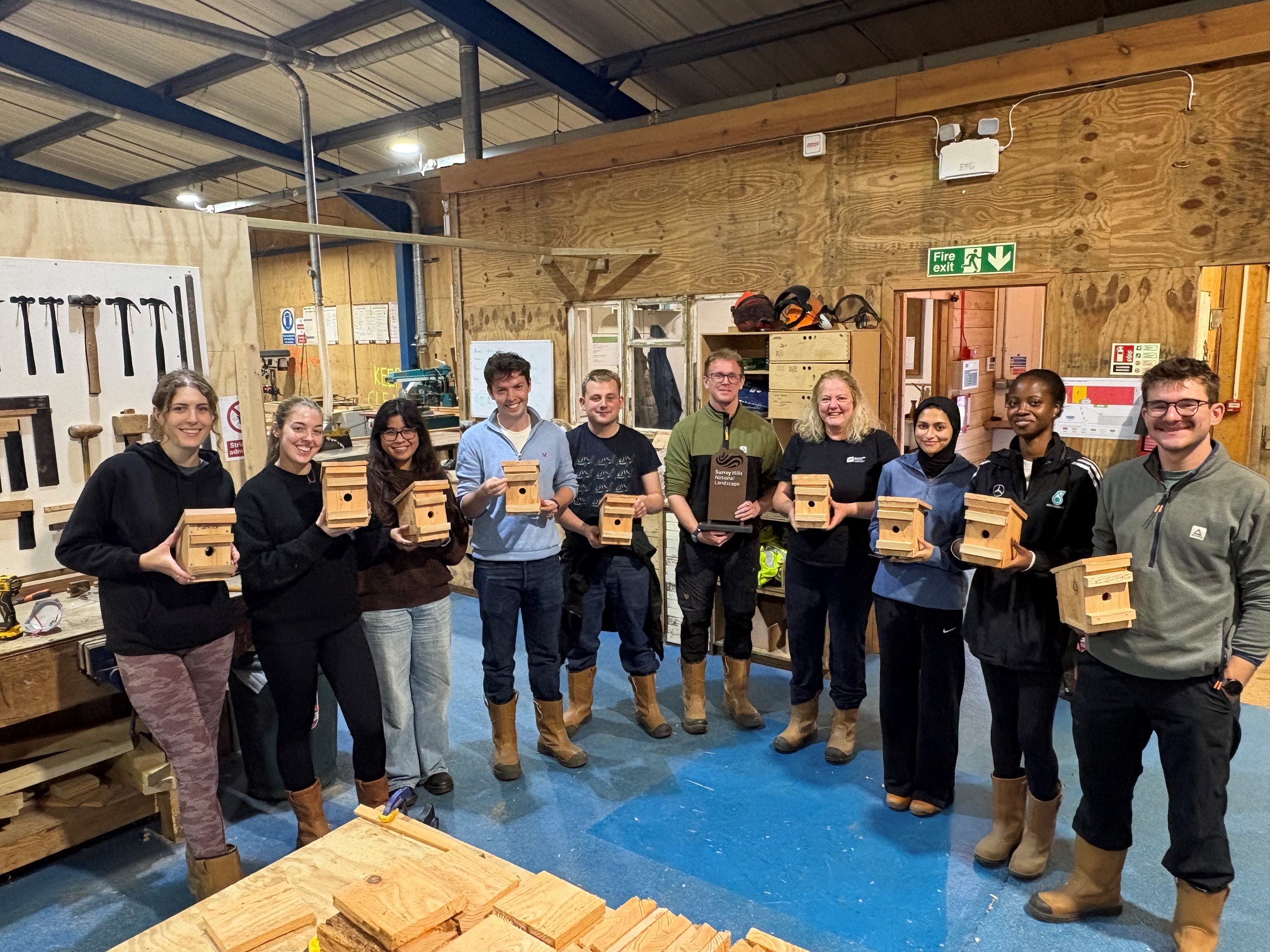 Ten people smiling at the camera, holding wooden dormouse boxes that they have created. All standing inside the sawmill.