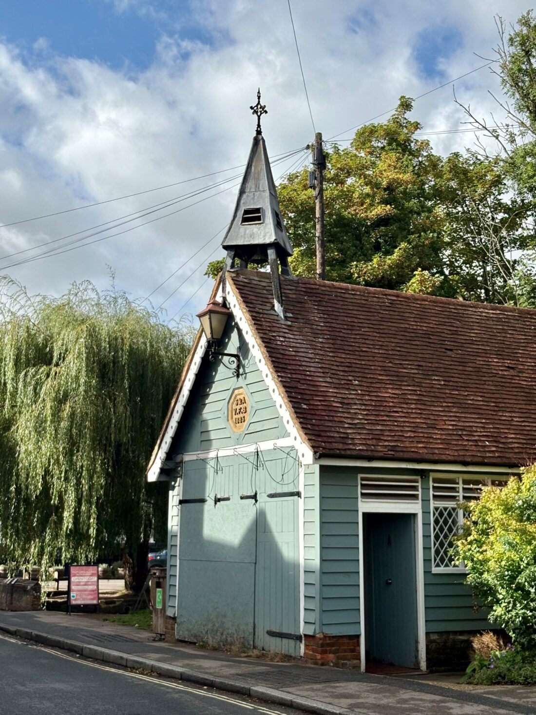Green-painted wooden building with steeple and brown-tiled roof. Blue sku with white clouds in the background and trees behind.